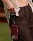 Leah apatite phone charm with evil eye charm attached to burgundy phone case, styled in hand against brown pants