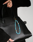 A model carries a black handbag adorned with The Leah Bag Charm in Apatite, showcasing its elegant design and protective symbolism through gold-plated Evil Eye charms and rich apatite quartz beads.
