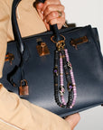 A model carries a black handbag featuring The Minou Bag Charm in Lavender, showcasing its stylish, protective design with lavender quartz beads, gold-plated details, and a bold Evil Eye charm.
