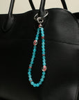 A black handbag adorned with The Leah Bag Charm in Turquoise, emphasizing its bright turquoise quartz beads and silver Evil Eye charms for a striking contrast.