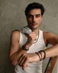 A man wearing the Ethan Wrap in 18K Gold as a bracelet, styled with a white tank top and a casual, relaxed pose, emphasizing the wrap’s versatile design.