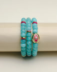 The Leah turquoise bracelet set displayed on a cylindrical stand, showcasing the bright blue-green beads with pink and gold detailing and an eye-catching central eye charm.