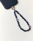 Flat lay of the Sapphire Leah Phone Charm attached to a black phone, showcasing deep-blue gemstone beads and rhodium-dipped evil eye accents.