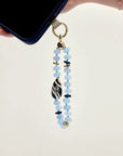 Mini Minou Aquamarine Quartz Phone Charm dangling from a dark phone case, highlighting the aqua beads and gold zebra evil eye charm.