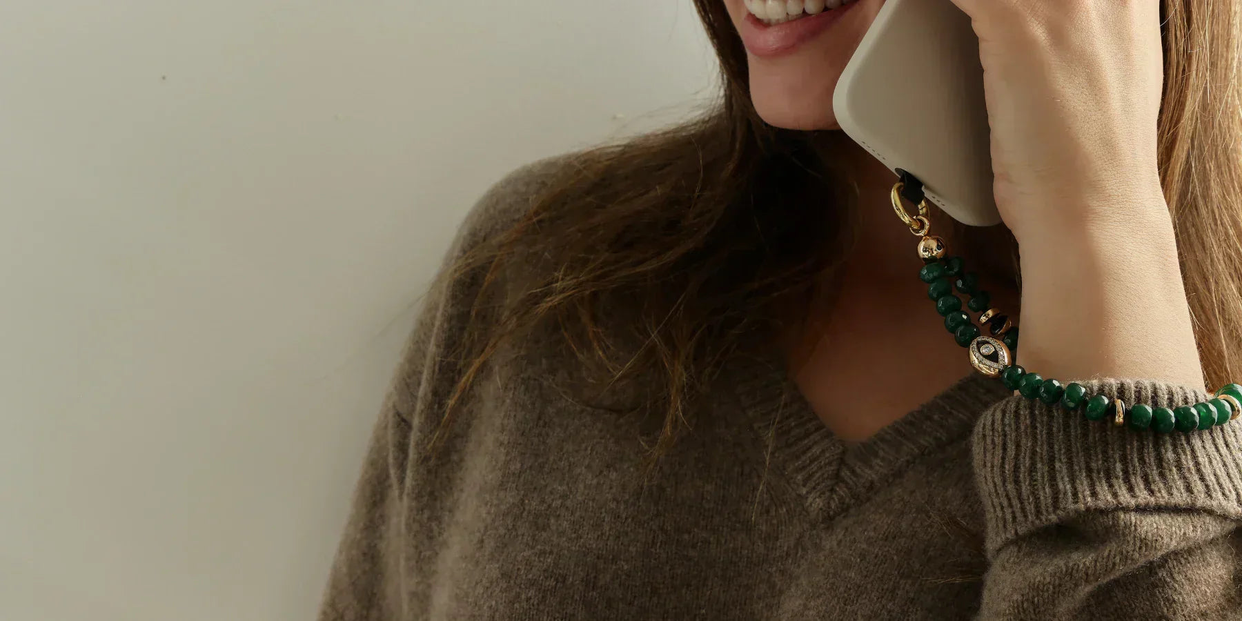 Leah phone charm in emerald quartz beads with gold evil eye charm attached to a beige phone case, shown worn on the wrist while holding a phone.