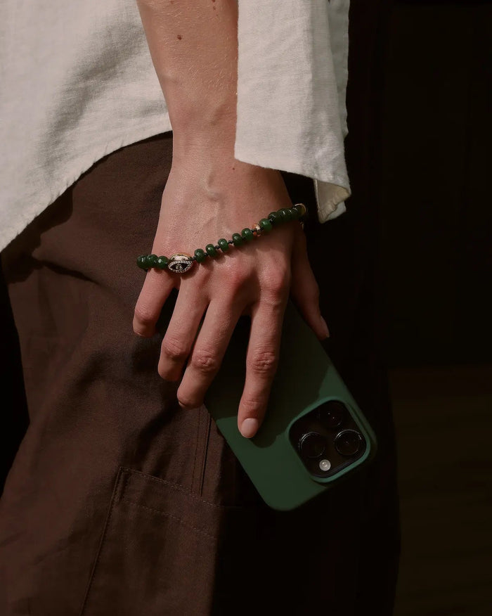 Leah emerald phone charm in gold wrapped around hand and attached to a dark green phone case against brown trousers