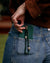 Leah emerald phone charm in gold hanging from a dark green phone case tucked into blue denim jeans