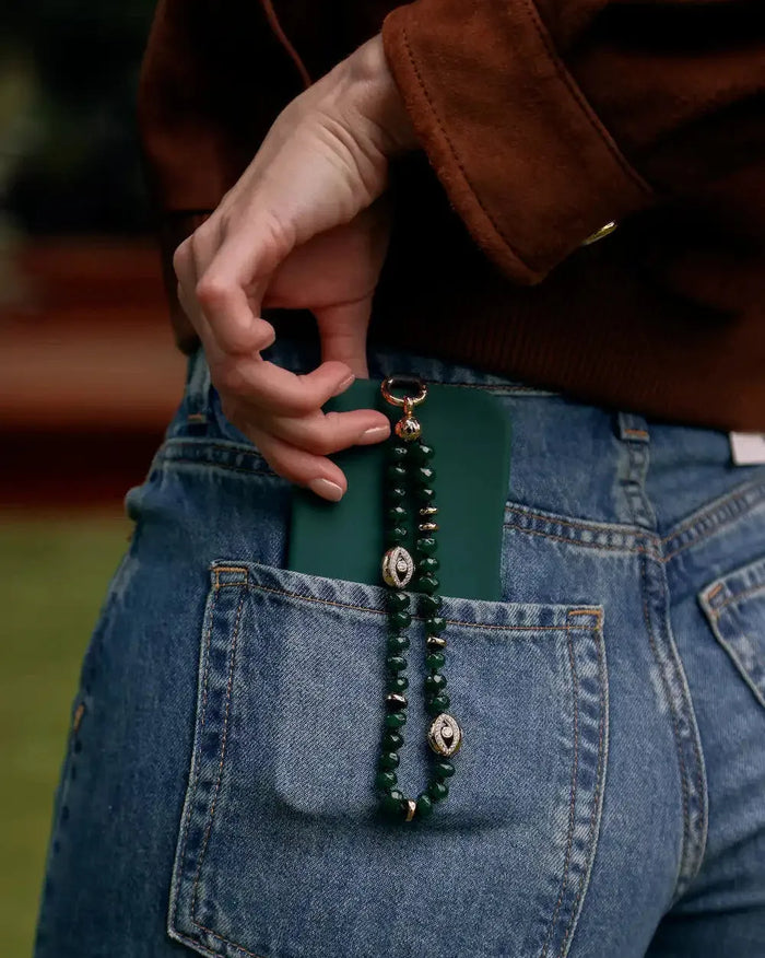 Leah emerald phone charm in gold hanging from a dark green phone case tucked into blue denim jeans