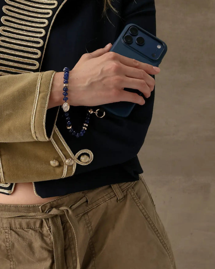 Leah sapphire phone charm styled around wrist while holding a navy phone case with black and gold military-inspired jacket