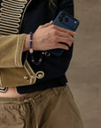 Leah sapphire phone charm styled around wrist while holding a navy phone case with black and gold military-inspired jacket