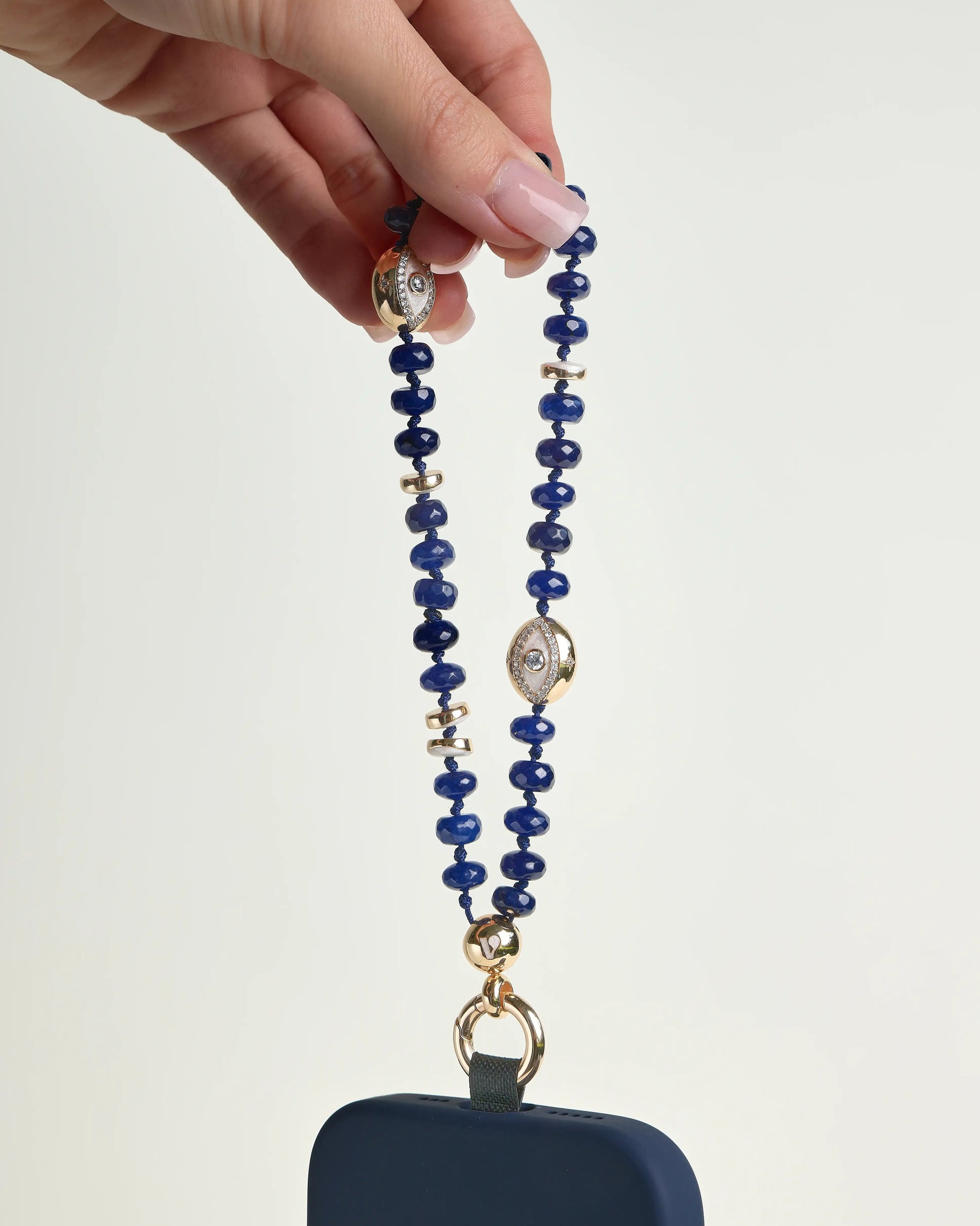 Leah sapphire phone charm with deep blue beads, gold accents, and evil eye charms attached to a navy phone case, held in hand against a white background