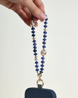 Leah sapphire phone charm with deep blue beads, gold accents, and evil eye charms attached to a navy phone case, held in hand against a white background