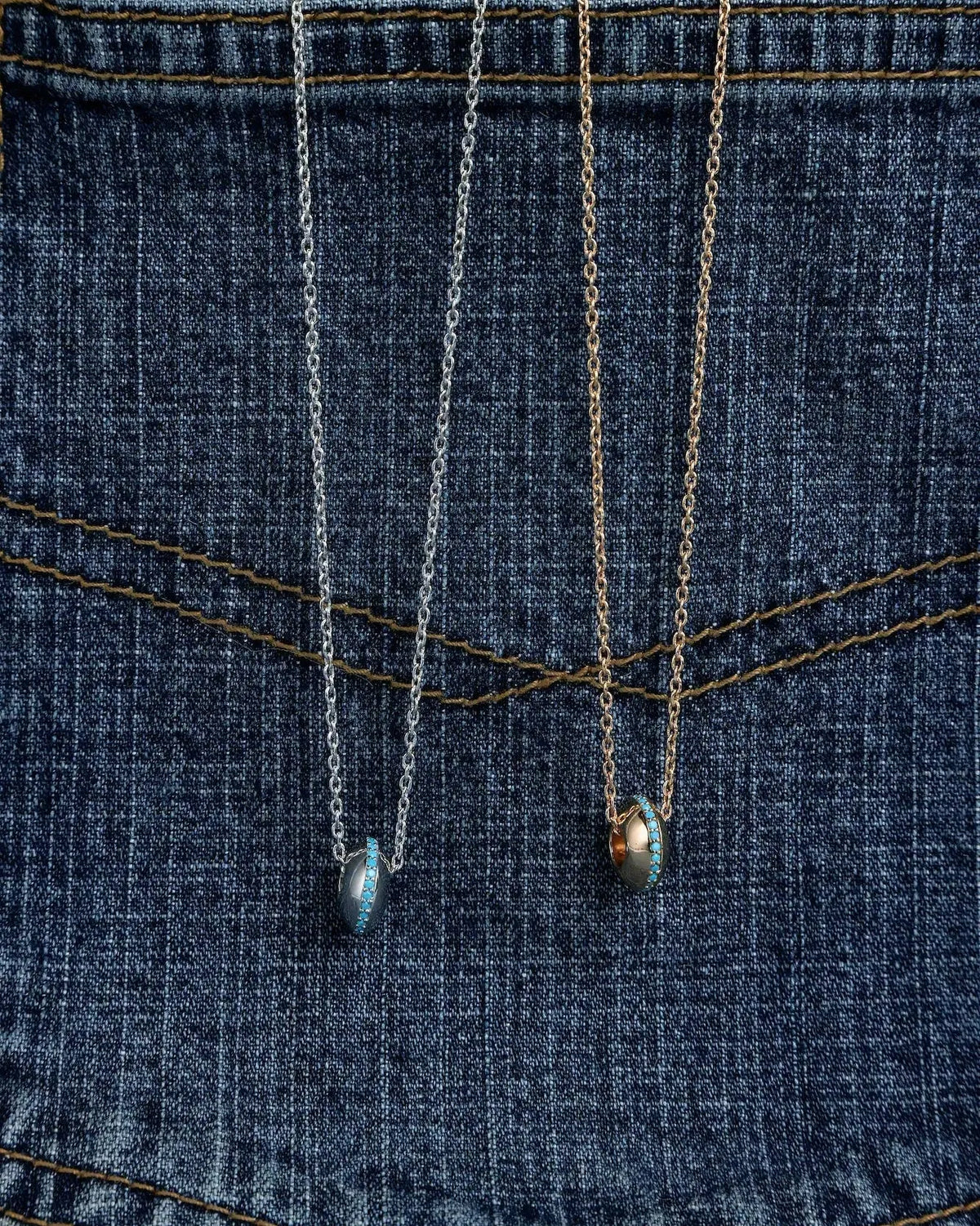 Two delicate Mia pendant necklaces in silver and gold finishes, draped over the back pocket of blue denim jeans.