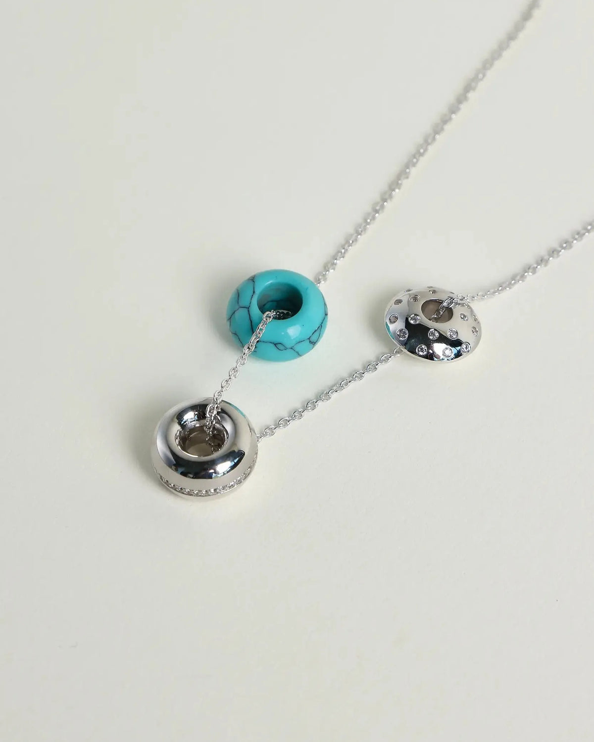 A silver-toned necklace with three hanging charms on a plain light background: one turquoise bead, one polished silver ring, and one textured silver ring with small dark inlays.