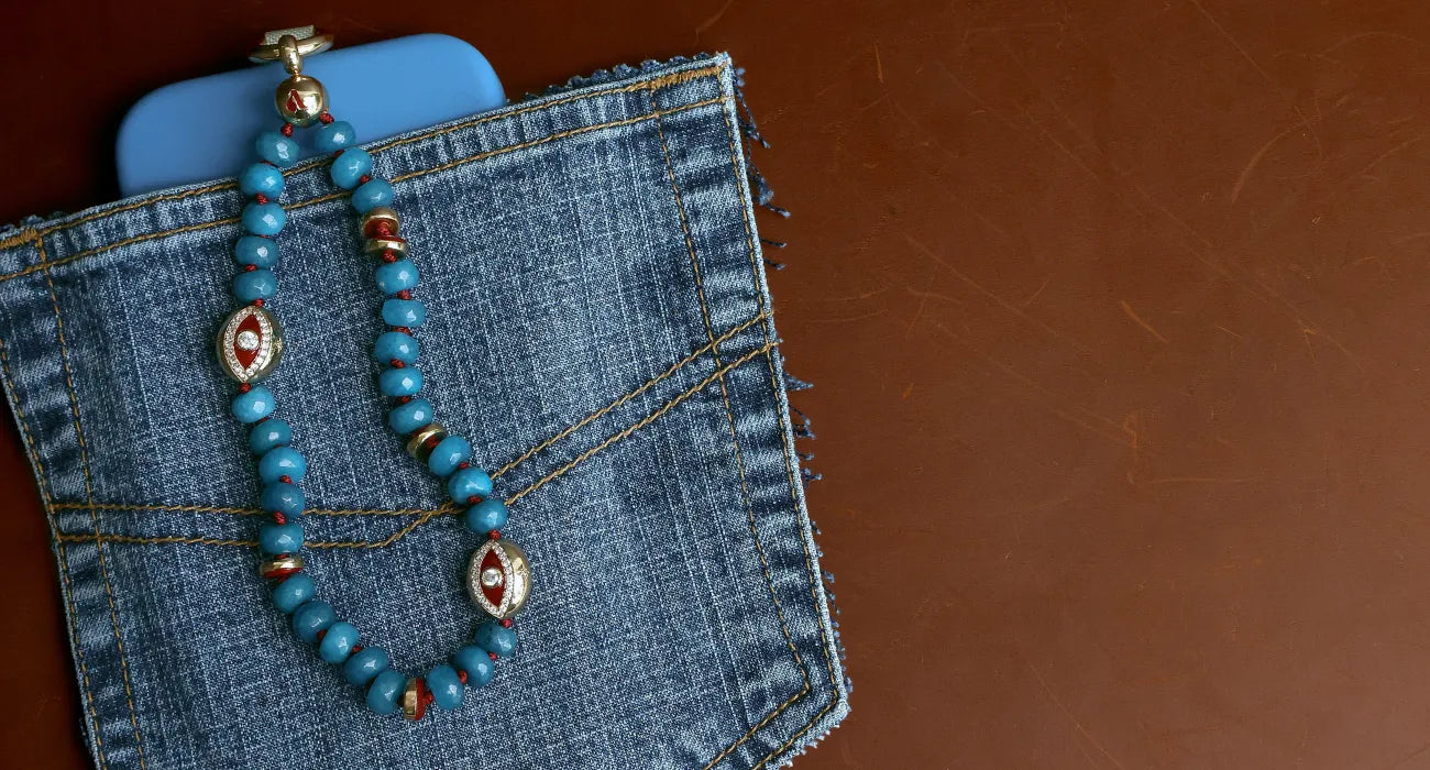 Leah phone charm in apatite with blue beads and gold evil eye charms attached to a light blue phone case, styled on a denim pocket flat lay