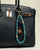 A black designer handbag with The Leah Bag Charm in Apatite attached to the handle. The charm features semi-precious apatite quartz beads, gold-plated Evil Eye charms, and luxe dividers, adding a refined and stylish touch.