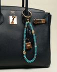A black designer handbag with The Leah Bag Charm in Apatite attached to the handle. The charm features semi-precious apatite quartz beads, gold-plated Evil Eye charms, and luxe dividers, adding a refined and stylish touch.