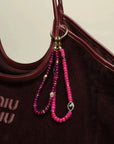 Leah Bag Charm in Amethyst & Stella Bag Charm in Pink attached to a burgundy designer tote – A stylish display of two bag charms, one in faceted amethyst quartz and the other in vibrant pink quartz, clipped onto a burgundy leather designer bag, showcasing their bold yet elegant accents.