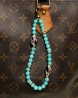 A brown designer handbag adorned with The Minou Bag Charm in Aqua, highlighting its vibrant aqua quartz beads and gold-plated Evil Eye charm for an elegant touch.