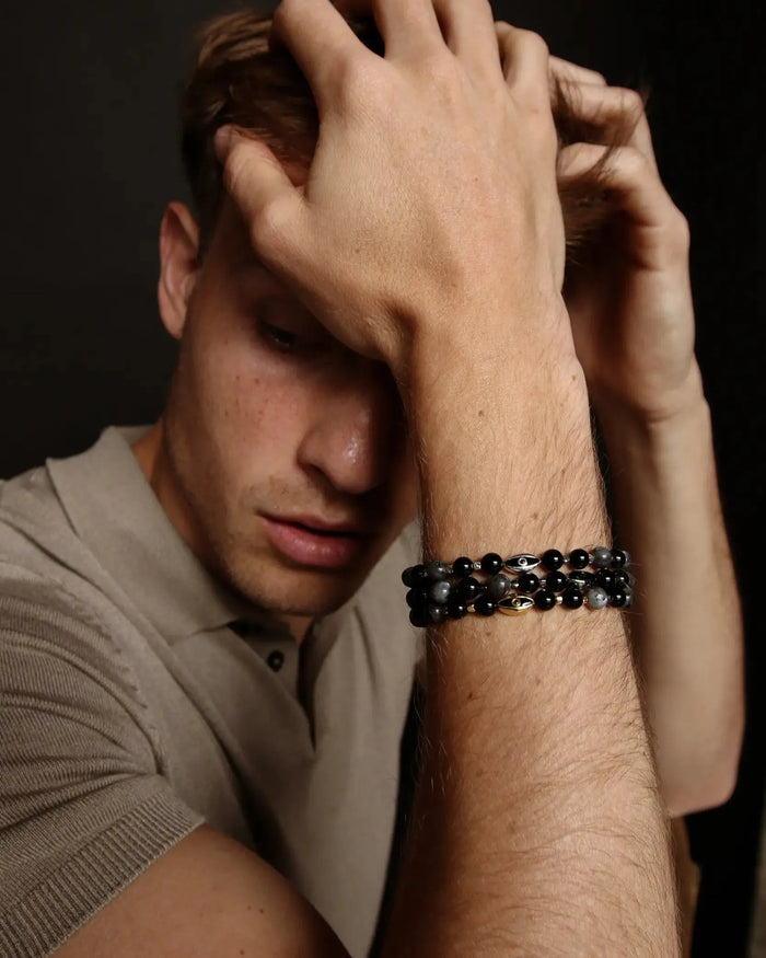 A man resting his hand on his head while wearing three David Bracelets in Black Rhodium, Gold, and Silver, emphasizing the contrast between the finishes and the understated, modern look.