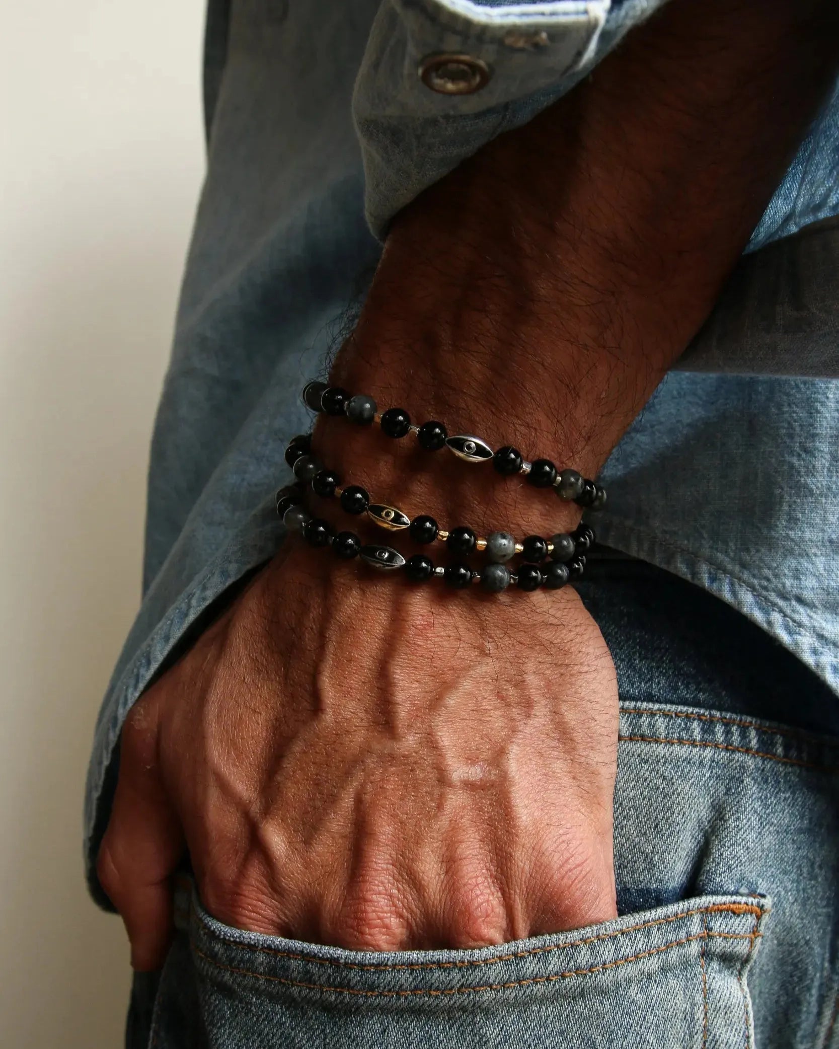 A man wearing multiple David Bracelets in Black Rhodium, Gold, and Silver, layered to showcase the bold matte black, polished gold, and silver finishes with black onyx and black labradorite quartz beads.