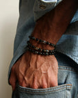 A man wearing multiple David Bracelets in Black Rhodium, Gold, and Silver, layered to showcase the bold matte black, polished gold, and silver finishes with black onyx and black labradorite quartz beads.