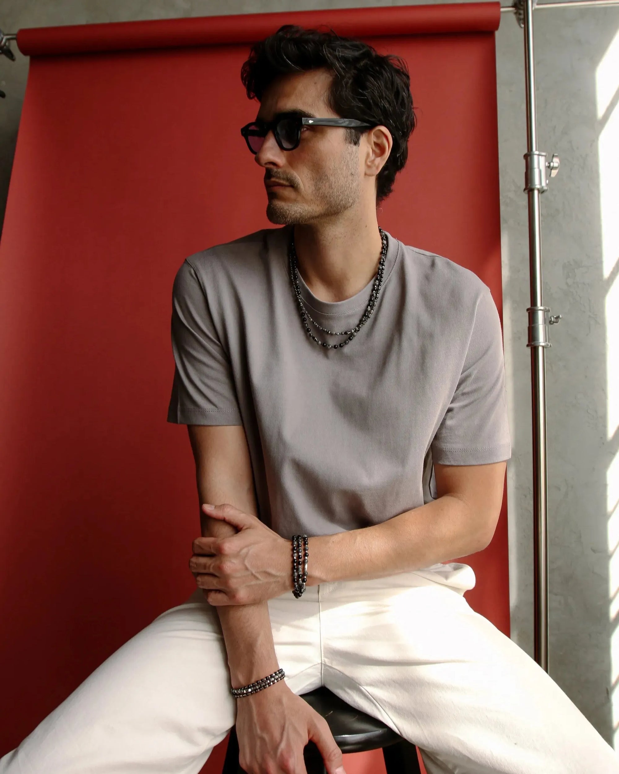 A man seated against a red backdrop, wearing the Ethan Wrap in Silver as a bracelet, styled casually with a relaxed, minimalist look.