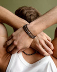 A man crossing his arms while wearing the Ethan Wrap in Silver as a bracelet and necklace, styled with a white tank top to showcase its versatile design and layered look.