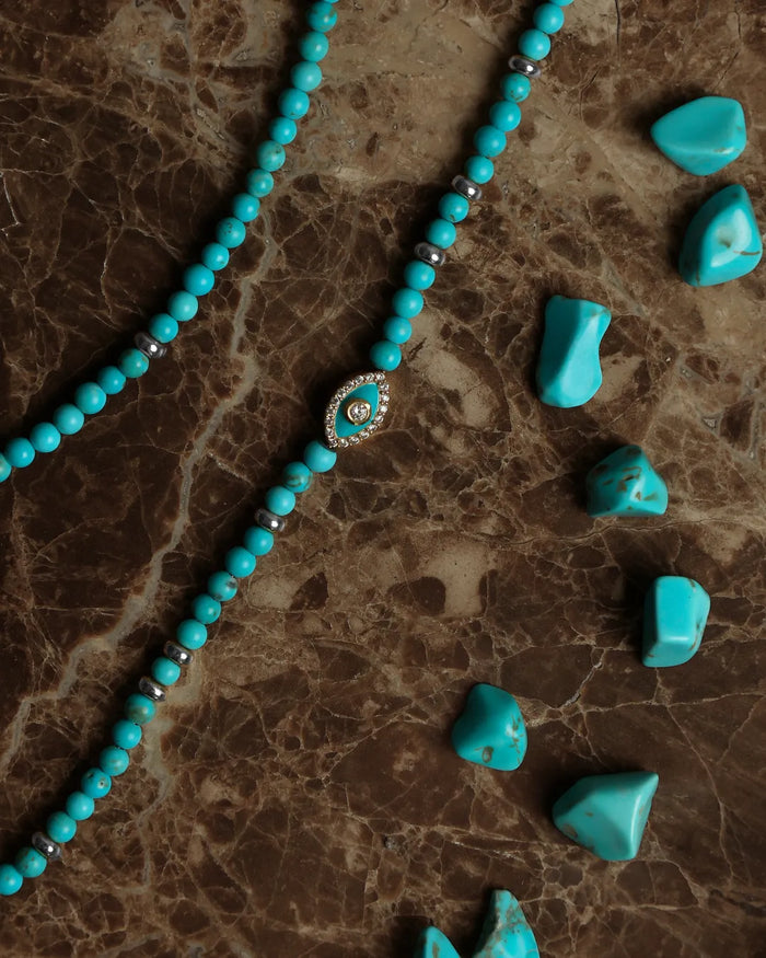 Turquoise beaded necklace with a silver evil eye charm, displayed on a brown marble surface alongside raw turquoise stone chips.