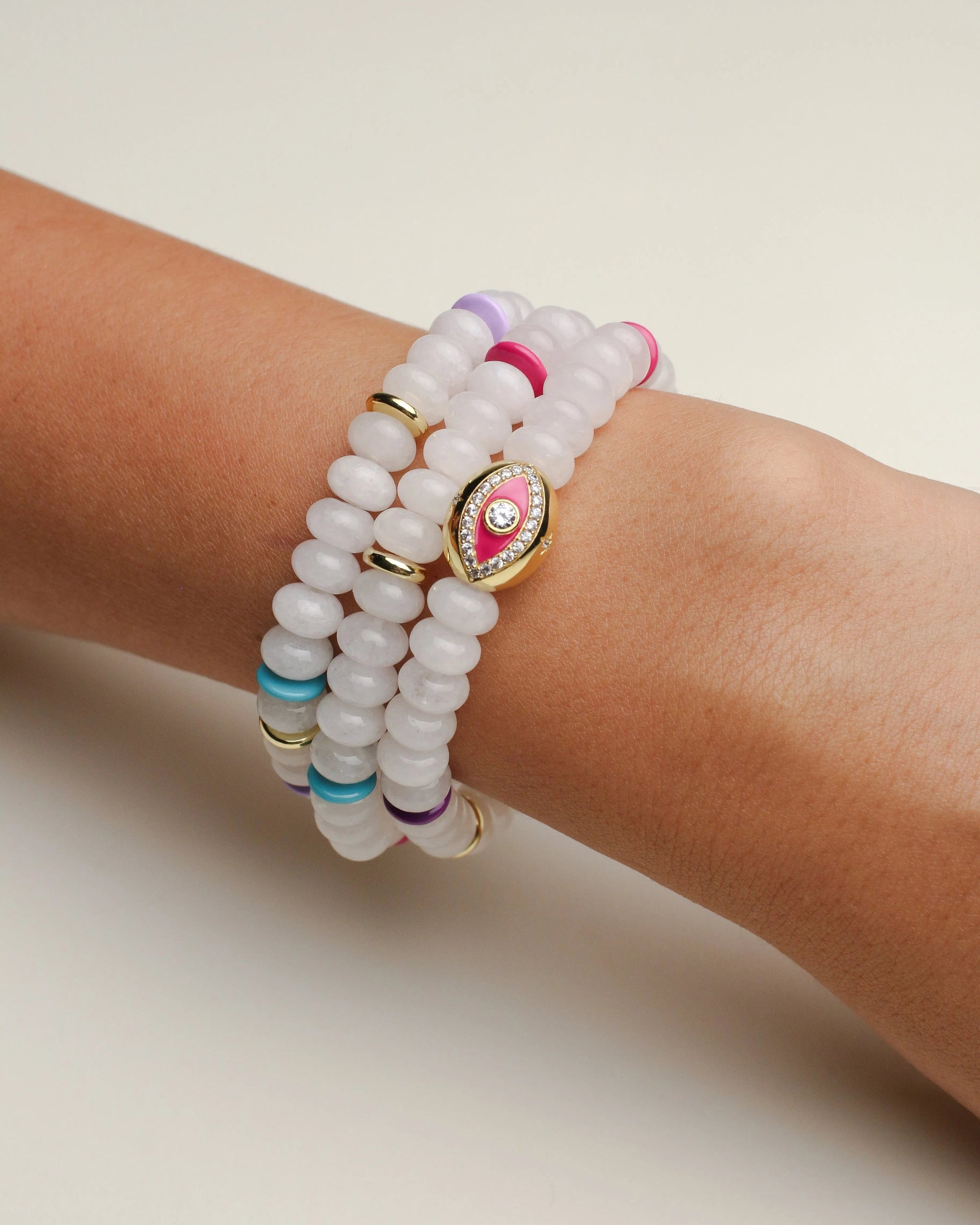 A set of three Leah white bracelets worn on the wrist, featuring milky white beads with colorful accents of pink, blue, and gold, highlighted by a central eye charm.