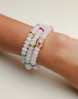 A set of three Leah white bracelets worn on the wrist, featuring milky white beads with colorful accents of pink, blue, and gold, highlighted by a central eye charm.