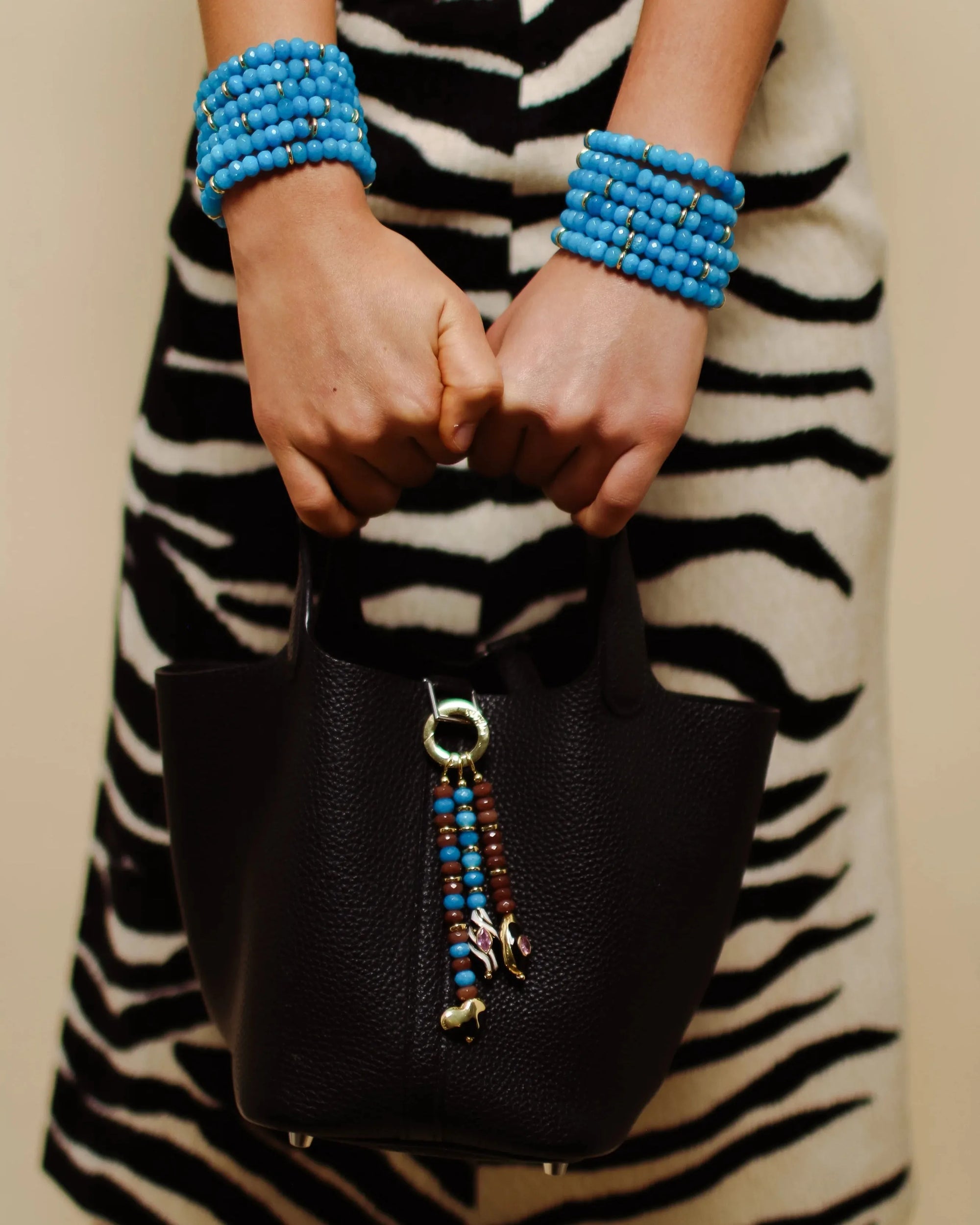 Close-up of the Liora Bag Charm in Azure &amp; Espresso, featuring a striking combination of blue and brown quartz beads accented with gold details, attached to a black leather bag. The charm adds a playful yet elegant touch to the bold zebra-patterned outfit.