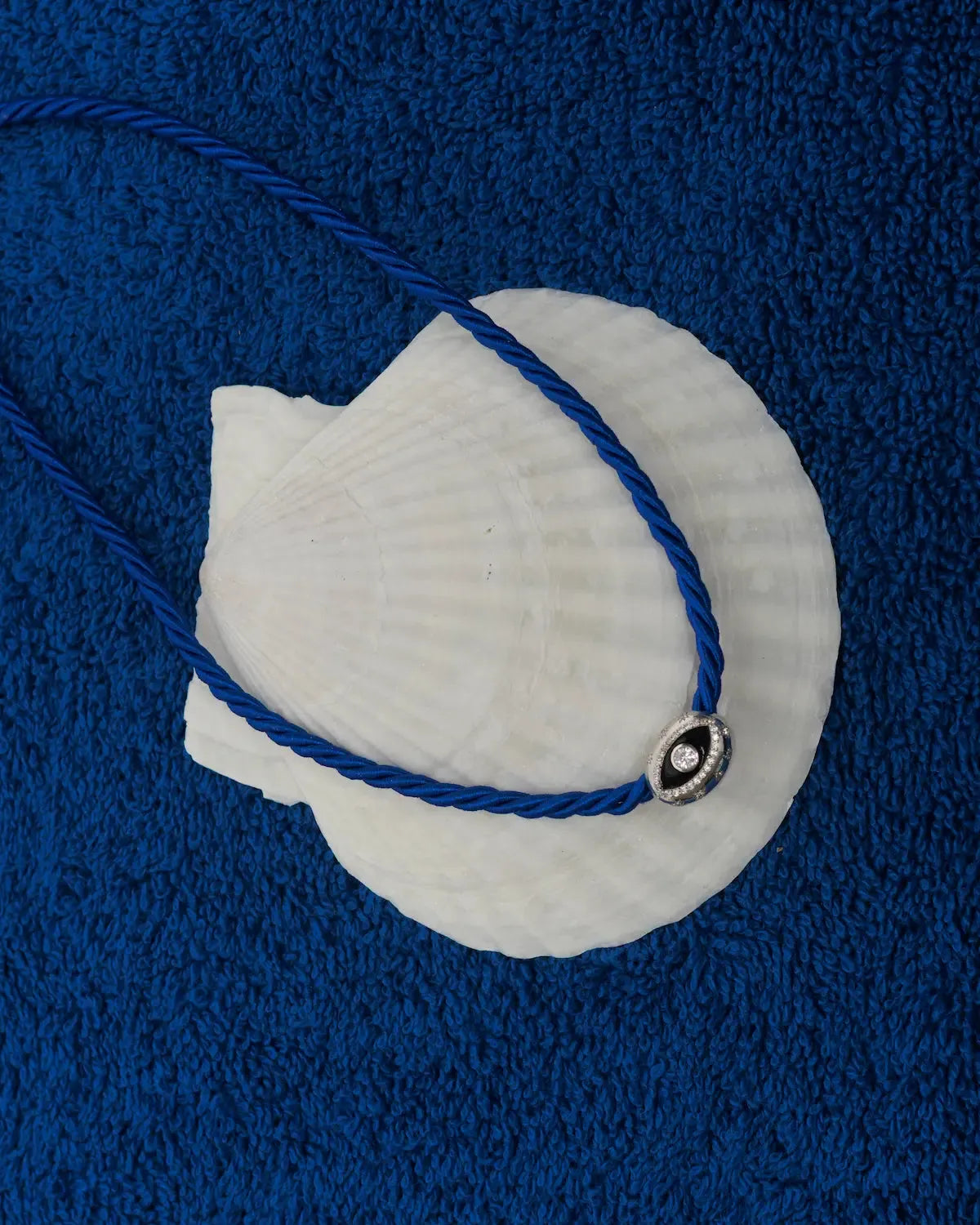 MyAleph Luna Cord Necklace in Azur with cobalt blue cord and silver evil eye pendant styled on seashell and blue textured background
