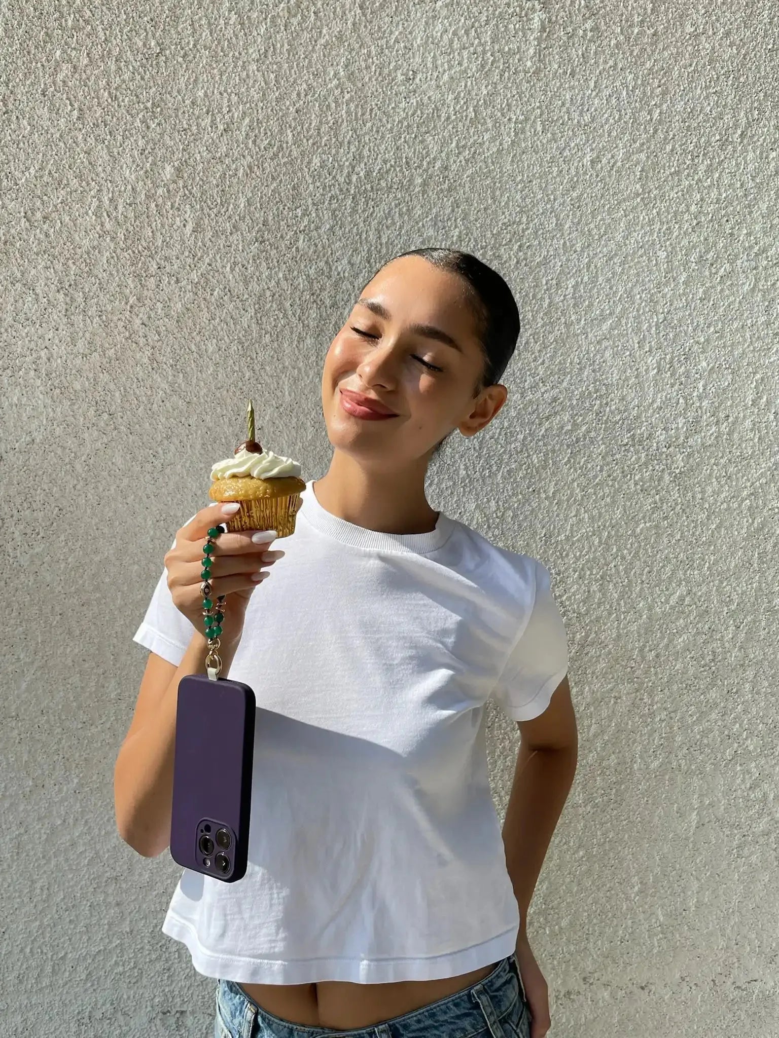 Woman wearing a white top holding a phone with the Mini Leah Emerald Quartz Phone Charm attached, in natural daylight.