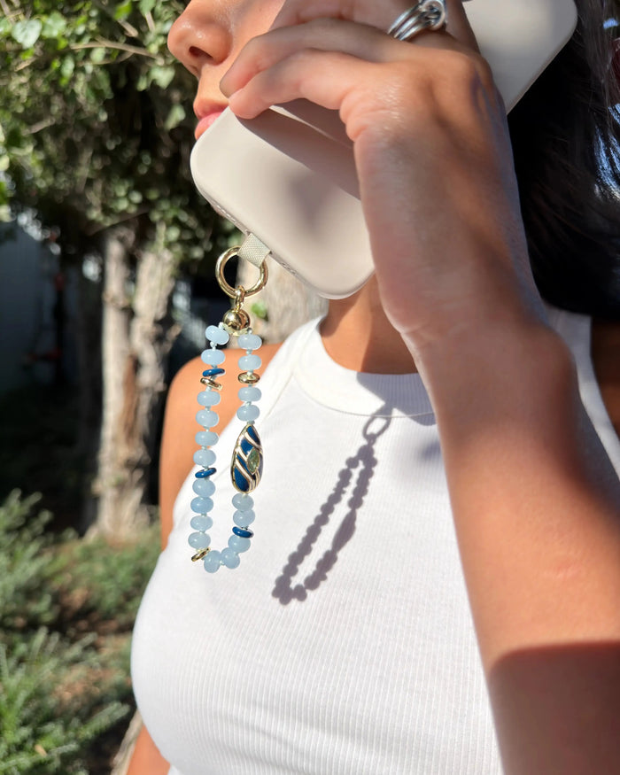 Close-up of a woman holding a white smartphone with a Mini Minou Aquamarine Phone Charm attached. The charm features light blue aquamarine beads with gold accents and a zebra evil eye talisman, catching the sunlight and casting a shadow. She wears a white ribbed tank top and silver rings.
