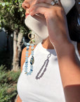 Close-up of a woman holding a white smartphone with a Mini Minou Aquamarine Phone Charm attached. The charm features light blue aquamarine beads with gold accents and a zebra evil eye talisman, catching the sunlight and casting a shadow. She wears a white ribbed tank top and silver rings.