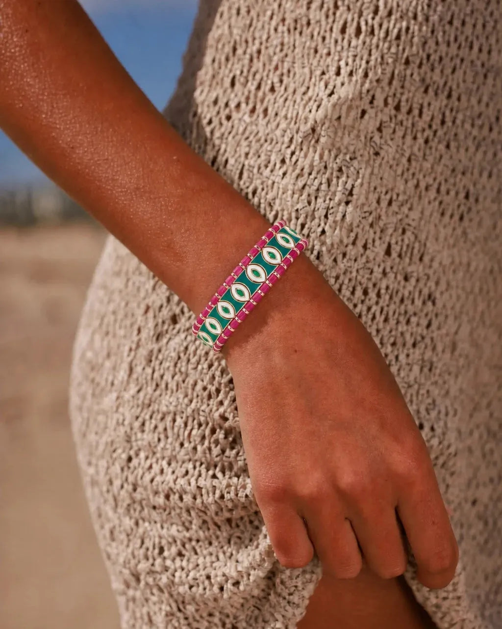 MyAleph Riviera Bracelet in Saint Tropez — emerald green and hot pink evil eye protective charm bracelet worn on wrist, styled with a cream knit summer dress on the beach.