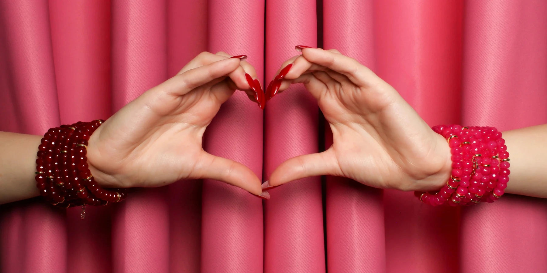 Two hands holding a red heart shape in front of pink curtains wearing ruby quartz and pink quartz bracelets.