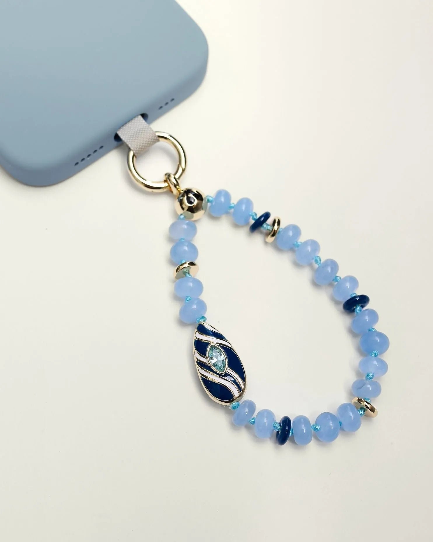 Mini Minou Aquamarine Quartz Phone Charm attached to a light blue phone case, featuring aqua-toned beads with a gold zebra evil eye charm.