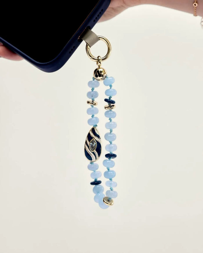 Mini Minou Aquamarine Quartz Phone Charm dangling from a dark phone case, highlighting the aqua beads and gold zebra evil eye charm.