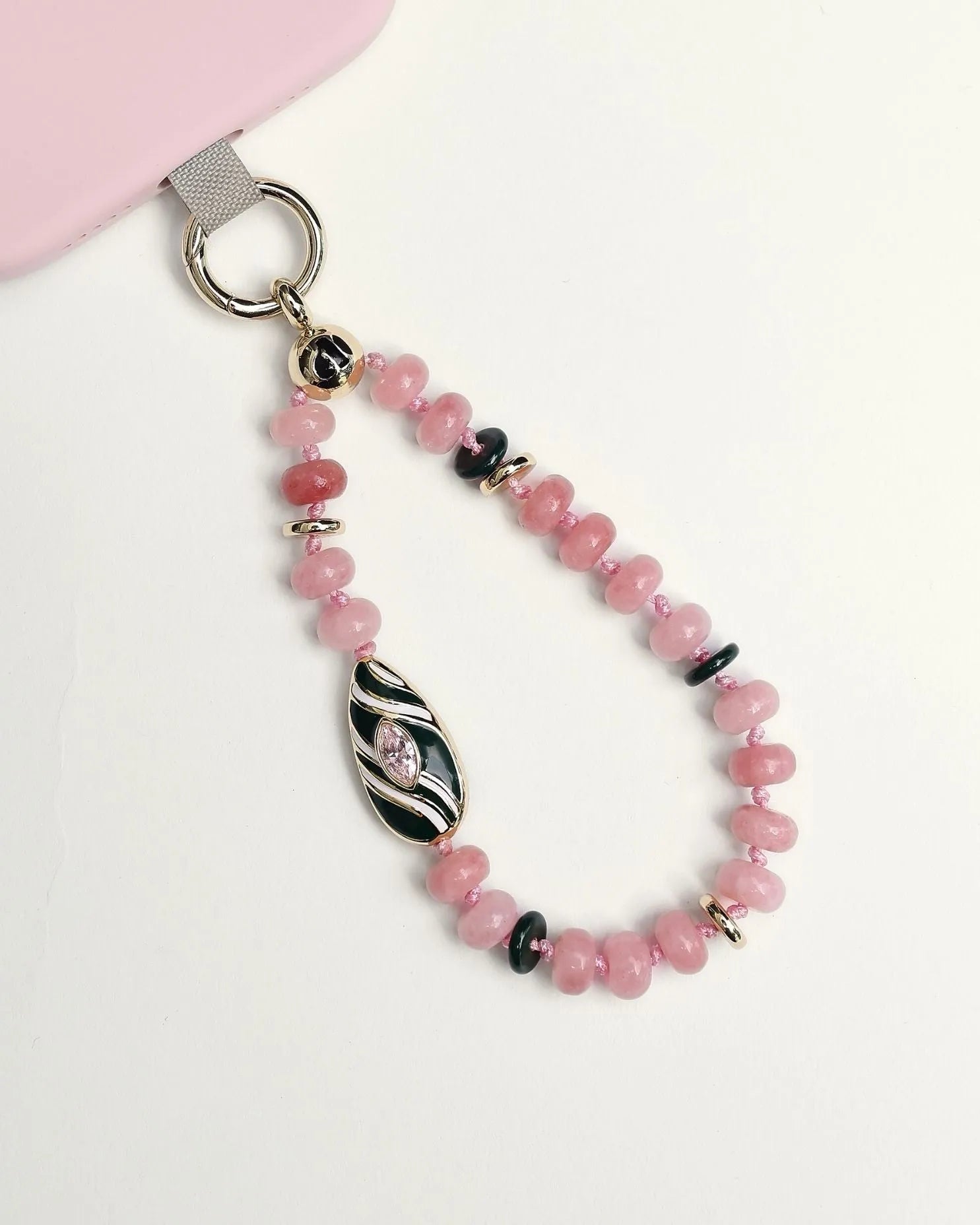 Mini Minou Rose Quartz Phone Charm attached to a pink phone case, showing pink quartz beads with a gold zebra evil eye charm with green enamel on a white background.